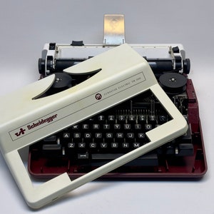 May include: A white electric typewriter with a red base. The brand name "Scheidegger" is printed on the front of the typewriter. The model name "ELECTROSTAR ELECTRIC TM 2000" is printed on the top of the typewriter.