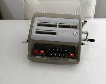 May include: A vintage gray Facit calculator with a silver handle and red buttons. The calculator has a display with numbers and a small yellow indicator.