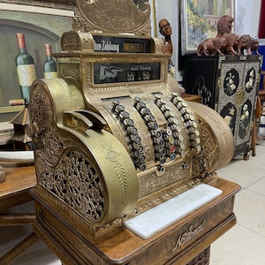 May include: An antique brass National cash register with ornate detailing. The register displays "AMOUNT PURCHASE" and "Mark Pfennig 55.50". It sits on a wooden base with a woven basket-like bottom.