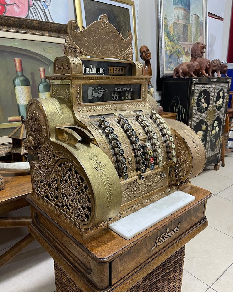 May include: An antique brass National cash register with ornate detailing. The register displays "AMOUNT PURCHASE" and "Mark Pfennig 55.50". It sits on a wooden base with a woven basket-like bottom.