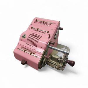 May include: A vintage pink mechanical calculator with multiple rows of number dials and a hand crank. The calculator has a metal frame with a series of levers and a small logo on the front. The color is a soft, pastel pink.