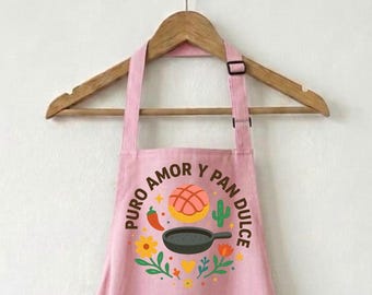 May include: A pink apron featuring the text "PURO AMOR Y PAN DULCE" in a circular design. The design includes a pan dulce pastry, a frying pan, a chilli pepper, and flowers. The apron is suspended from a wooden hanger.