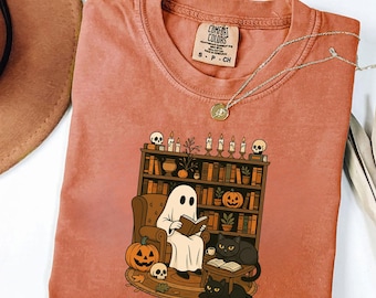 Puede incluir: Camiseta Comfort Colors de color terracota con un diseño de Halloween. El diseño muestra un fantasma leyendo en una silla frente a una estantería, con calabazas, calaveras y gatos negros. También se ve un collar plateado con un colgante.