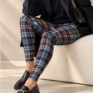 May include: A navy blue wool coat, a black ribbed turtleneck jumper, and blue plaid leggings with black, white, and yellow lines. The outfit includes a black shoulder bag with a gold chain strap and black leather loafers with gold hardware.