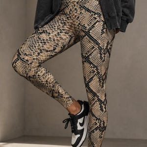 May include: High-waisted leggings featuring a tan and black snakeskin print pattern. The leggings are paired with a white cropped tank top, a charcoal grey zip-up hooded sweatshirt, and black and white low-top athletic sneakers with laces.