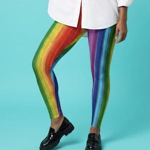 May include: A person wearing a white button-down shirt and rainbow-striped leggings. The leggings feature vertical stripes in all the colors of the rainbow. The person is also wearing black loafers.