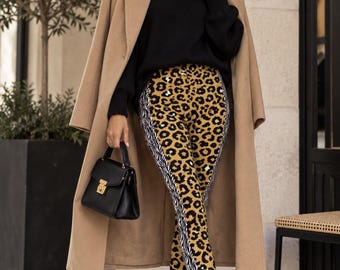 May include: A camel-colored wool coat worn over a black turtleneck sweater and leggings featuring a central leopard print panel flanked by vertical black and white zebra print stripes, paired with black loafers and a black handbag.