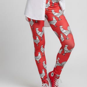 Pode incluir: Leggings vermelhas com um padrão repetido de galos em preto e branco. As leggings são combinadas com tênis vermelhos. A roupa é completada com uma camisa branca.