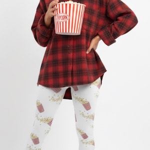 Puede incluir: Una persona con una camisa de franela a cuadros rojos y negros y leggings blancos con un estampado de palomitas de maíz. Sostiene un cubo de palomitas de maíz. El atuendo es casual y cómodo, ideal para relajarse.