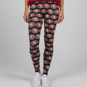 May include: Black leggings featuring an all-over repeating pattern of the San Francisco 49ers football team logo. The logo consists of a red oval with a gold border and the letters SF in white. The leggings are paired with plain white sneakers.