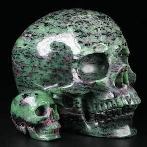 May include: Two green, pink, and black gemstone skulls carved from zoisite. The larger skull is about 15 cm tall and the smaller skull is about 5 cm tall.