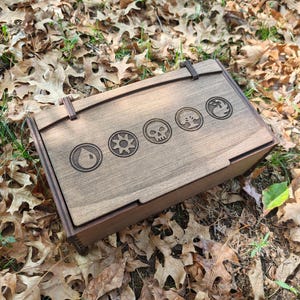 May include: A rectangular dark brown wooden box with a hinged lid. The top surface features five circular laser-engraved symbols representing a water drop, a sun, a skull, a tree, and a flame, arranged in a horizontal row across the center.