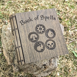 Puede incluir: Un libro de madera con el título "Book of Spells" grabado en la portada. La portada presenta cinco símbolos circulares. El libro descansa sobre una roca, con hierba en el fondo. El libro es de madera oscura.