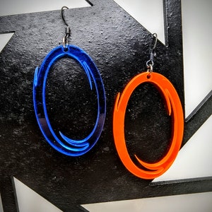 May include: A pair of acrylic earrings, one blue and one orange, each shaped like an oval with a curved design. The earrings hang from black ear wires. The background is a black geometric shape.