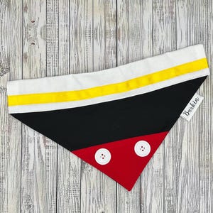 Puede incluir: Un pañuelo para perros con un diseño negro, blanco, amarillo y rojo. El pañuelo tiene una parte superior blanca con una raya amarilla, un centro negro y una parte inferior roja con dos botones blancos. La palabra "Barkin" está impresa en el lateral.