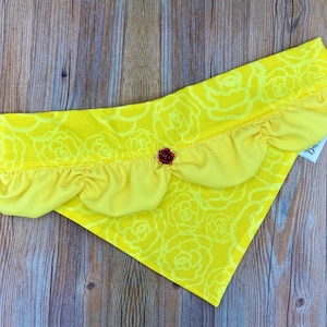 Könnte beinhalten: Gelber Stoffhundebandana mit einem Rüschenrand und einer roten Rosenverzierung. Das Bandana hat ein weißes Rosenmuster.