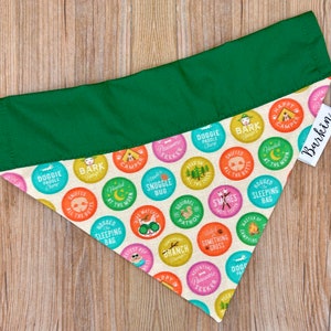 Puede incluir: Un pañuelo para perros con la parte superior verde y la inferior estampada. La parte inferior presenta círculos coloridos con texto e ilustraciones con temática de camping. Se adjunta una etiqueta blanca con la palabra "Barkin".