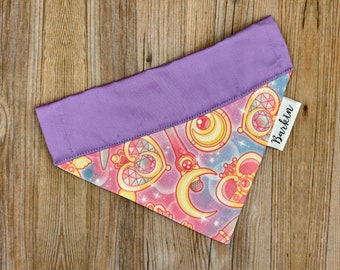 May include: A triangular pet bandana featuring a solid purple top band and a patterned lower section with yellow crescent moons, pink hearts, and star motifs on a blue and pink background. A white fabric tag with the text Barkin is attached.