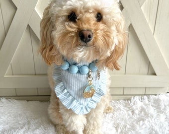 May include: A small, light brown dog wearing a blue and white striped bandana with a ruffled edge. The bandana has a blue beaded necklace and a gold charm attached. The dog is wearing a white bow on its head and is sitting on a white fluffy surface.