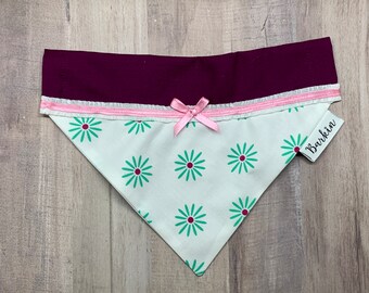 May include: A triangular pet bandana featuring a white fabric base with a repeating pattern of teal daisies with dark pink centers. The top section is a solid deep burgundy fabric, accented by a thin white ruffled trim, a pink ribbon bow, and a white fabric tag with the word Barkin printed in black.