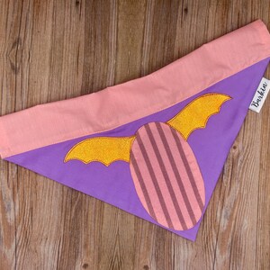 May include: A triangular pet bandana featuring a purple base with a pink top border. The center displays an oval patch with vertical pink stripes and two glittery gold wings extending from the sides. A white fabric tag reads Barkin.