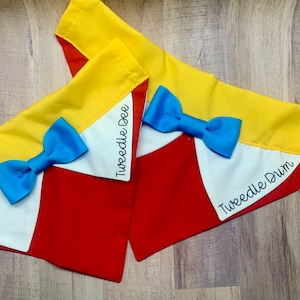 May include: Two triangular pet bandanas featuring a yellow top border, a white center section, and a red bottom. Each bandana includes a bright blue bow tie attached to the front. One bandana has the text Tweedle Dee, the other Tweedle Dum.