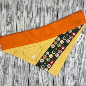 May include: A dog bandana with a yellow and orange design. The bandana has a pocket with a button and a patterned strip with various outdoor-themed icons. The tag reads "Barkin".