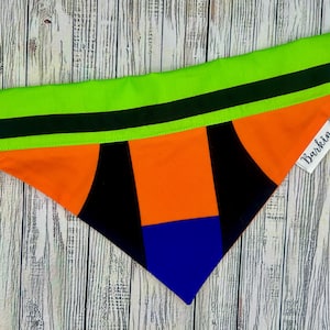 Puede incluir: Un pañuelo para perros con un diseño geométrico colorido. El borde superior es de color verde brillante con una franja negra, que pasa a secciones naranjas, negras y azules. La etiqueta dice "Barkin".