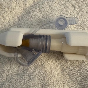 May include: A white plastic medical device with a clear plastic tube and a purple and yellow connector. The device is designed to hold a medical tube in place.