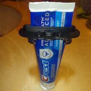 Puede incluir: Un dispensador de pasta de dientes de plástico negro con un tubo de pasta de dientes Crest azul y blanco. El dispensador tiene una palanca que empuja la pasta de dientes fuera del tubo. El tubo dice "Crest" y "Pro-Health Advanced" y "Deep Clean" y "Sensibilidad, Placa, Sarro". El tubo también dice "1 Menta" y "Refresca el aliento".