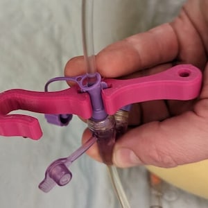 May include: A pink plastic medical device with a hook and a purple connector. The device is used to secure a medical tube.