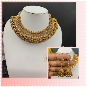 Op de afbeelding: Een gouden ketting met witte parel accenten en bijpassende oorbellen. De ketting heeft een gelaagd ontwerp met meerdere rijen kralen en een centrale hanger. De oorbellen zijn van het hangende type met een enkele parel en een belvormige hanger.