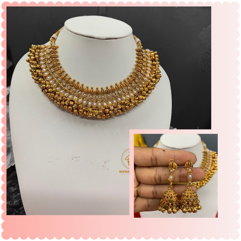 Op de afbeelding: Een gouden ketting met witte parel accenten en bijpassende oorbellen. De ketting heeft een gelaagd ontwerp met meerdere rijen kralen en een centrale hanger. De oorbellen zijn van het hangende type met een enkele parel en een belvormige hanger.
