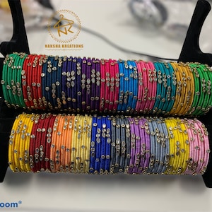 May include: A stack of colorful silk thread bangles with small, clear, oval-shaped embellishments. The bangles are arranged in a rainbow pattern on a black display stand. The text "NAKSHA KREATIONS" is visible on the stand.