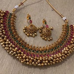 Op de afbeelding: Een gouden ketting met roze en groene stenen en een bijpassend paar oorbellen. De ketting heeft een rij kleine gouden balletjes langs de onderkant.