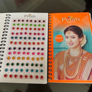 May include: A book with a white cover and the word "Petals" in red. The book has a spiral binding and is filled with pages of colorful round stickers with rhinestones. The book also has a picture of a woman wearing a gold necklace and earrings.