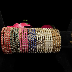 May include: A set of 12 gold bangles with square-shaped stones. The bangles are in various colors, including red, green, pink, blue, and clear. The bangles are arranged in a row on a black surface.