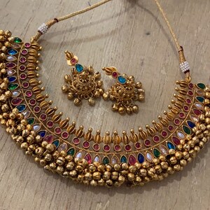 Op de afbeelding: Een gouden ketting met een veelkleurig stenen ontwerp en een bijpassend paar oorbellen. De ketting heeft een rij kleine gouden balletjes langs de onderkant.