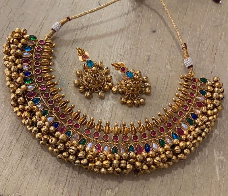 Op de afbeelding: Een gouden ketting met een veelkleurig stenen ontwerp en een bijpassend paar oorbellen. De ketting heeft een rij kleine gouden balletjes langs de onderkant.
