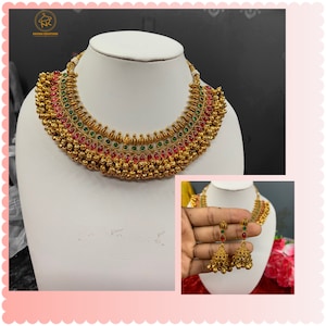 Op de afbeelding: Een gouden ketting- en oorsteensets met rode en groene stenen. De ketting heeft een gelaagd ontwerp met meerdere rijen gouden kralen en stenen. De oorbellen zijn van het hangende type met een klokvormig ontwerp.