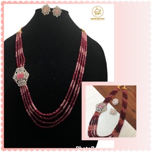 May include: A multi-strand necklace with pink and burgundy beads and a large, square-shaped pendant with a pink stone and clear rhinestones. The necklace is displayed on a black mannequin bust. A pair of matching earrings with pink stones and clear rhinestones are also shown.