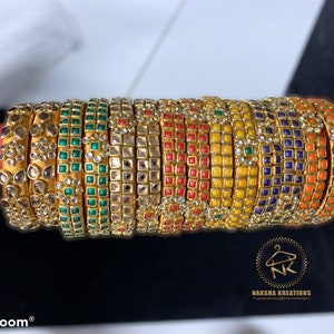 May include: A stack of 12 gold bangles with colorful enamel and rhinestone accents. The bangles are arranged in a circular pattern, with the gold color dominating the image. The bangles are decorated with various colors, including red, yellow, green, blue, and orange. The bangles are adorned with rhinestones and enamel accents, giving them a luxurious and elegant look.