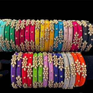 May include: A collection of colorful bangles, each wrapped in vibrant silk thread. The bangles are adorned with gold-colored floral embellishments and small, round beads. The color palette includes shades of green, pink, yellow, and blue.