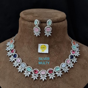 May include: A silver necklace and earrings set with a multi-colored stone design. The necklace features a row of oval-shaped stones in various colors, including pink, blue, green, and red, surrounded by smaller clear stones. The earrings have a similar design with a single oval-shaped stone and smaller clear stones.