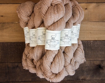 Könnte beinhalten: Mehrere Stränge hellbrauner Wolle, beschriftet mit "Mother Nature's Finest Fibers USA Alpaca". Die Wolle hat eine natürliche, weiche Farbe, und die Etiketten zeigen eine Alpaka-Zeichnung. Die Stränge sind auf einer Holzoberfläche angeordnet.