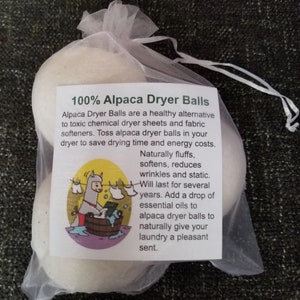 May include: A transparent mesh bag filled with several off-white alpaca dryer balls. The attached tag reads "100% Alpaca Dryer Balls" and details their benefits. The tag also features a cartoon of an alpaca doing laundry. The balls are a natural alternative to dryer sheets.