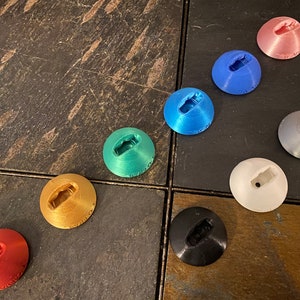 May include: Assortment of ten small, round, colorful, 3D printed objects with a small hole in the center. The objects are arranged in a semi-circle on a wooden surface.