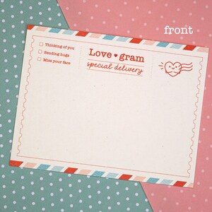 May include: A blank, cream-colored postcard with a red and blue border. The postcard has a red heart with a smiling face and the text "Love gram special delivery" on the top right corner. The text "Thinking of you", "Sending hugs", and "Miss your face" is written in a checklist format on the left side of the postcard.