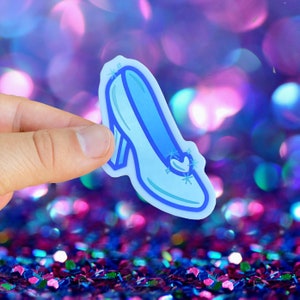 May include: A blue glass slipper with a pointed toe and a small heart-shaped design on the top. The slipper has a sparkly design and is outlined in a darker blue.