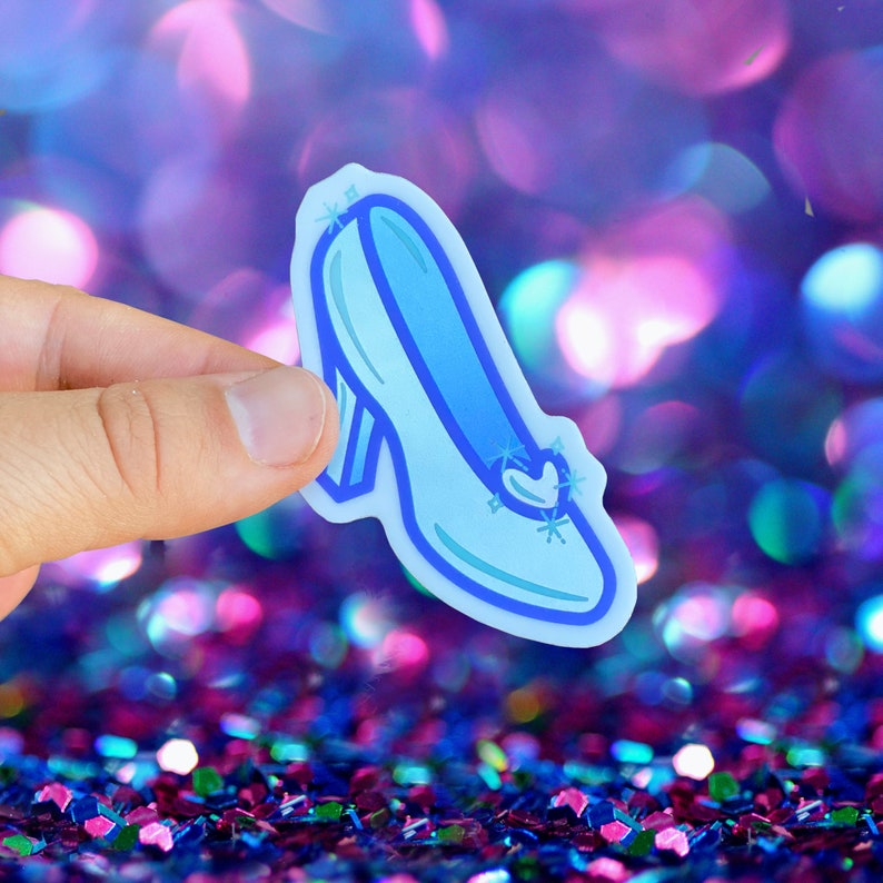 May include: A blue glass slipper with a pointed toe and a small heart-shaped design on the top. The slipper has a sparkly design and is outlined in a darker blue.